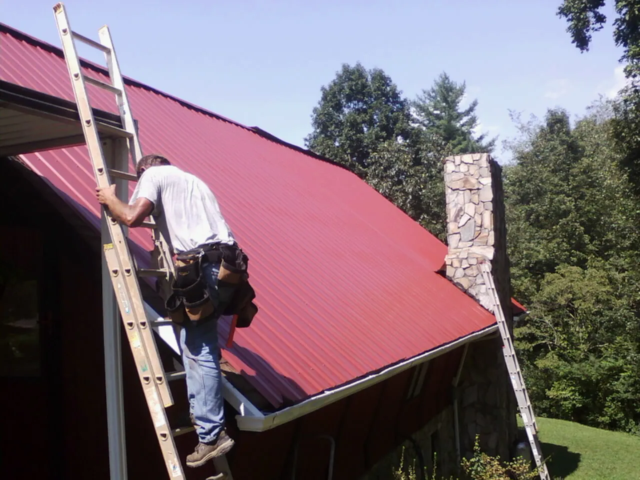 Roofer on a ladder beside a red metal roof and stone chimney — Soffit & Fascia Repair by Roofing Brothers in Silver Springs